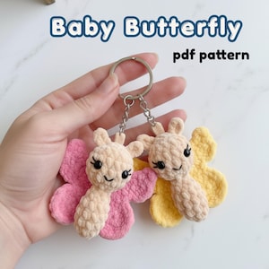 May include: Two crocheted butterfly keychains made from soft beige yarn with pink and yellow wings. Each butterfly features black embroidered eyes and antennae, attached to a silver metal keyring. The text Baby Butterfly pdf pattern is visible.