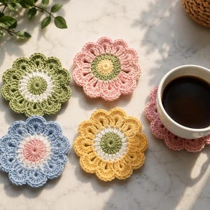 May include: A set of five round, flower-shaped crochet coasters in various colors including sage green, soft pink, light blue, and yellow. Each coaster features a central circular design with radiating petals. One pink coaster supports a white ceramic mug.