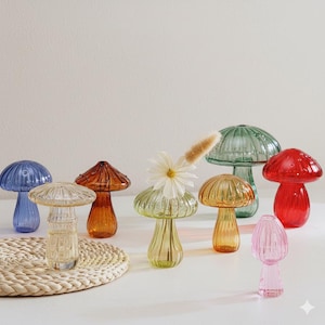 Könnte beinhalten: Eine Sammlung farbenfroher, pilzförmiger Glasvasen. Die Vasen sind in verschiedenen Farben erhältlich, darunter Blau, Orange, Grün, Gelb und Rot. Eine Vase enthält eine weiße Gänseblümchen und eine Trockenblume. Die Vasen stehen auf einer weißen Oberfläche.
