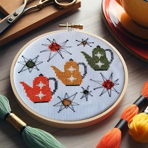 Może przedstawiać: Białe tamborek do haftu z haftem krzyżykowym przedstawiającym trzy kolorowe czajniki i stylizowane gwiazdy. Czajniki są pomarańczowe, żółte i zielone. Tamborek leży na drewnianej powierzchni z pomarańczową, żółtą i zieloną nicią do haftu.