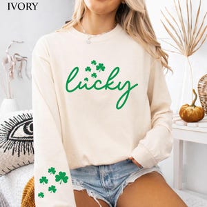 May include: Ivory long-sleeve sweatshirt with the word "lucky" in green script, accompanied by green shamrock graphics. Additional shamrocks are on the left sleeve. The sweatshirt is paired with denim shorts.