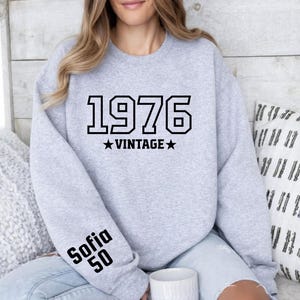 May include: A light gray sweatshirt with the year "1976" in bold black letters, with "VINTAGE" and two stars below. The left sleeve has the name "Sofia" and the number "50" printed in black. The sweatshirt is a casual style.