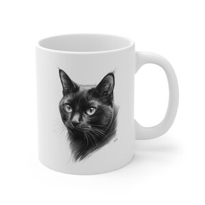 Op de afbeelding: Witte keramische mok met een gedetailleerde zwart-wit illustratie van het gezicht van een zwarte kat. De ogen van de kat zijn open en de snorharen zijn zichtbaar. De mok heeft een standaard handvat.