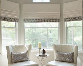 May include: Two white upholstered chairs with silver sequin pillows sit in front of a bay window with white roman shades and brown trim. A small metal table with a glass top sits between the chairs.