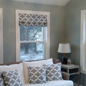Peut inclure: Un salon avec un canap&eacute; blanc et des coussins &agrave; motifs gris. Il y a trois fen&ecirc;tres avec des encadrements blancs et des stores romains &agrave; motifs gris. Une lampe se trouve sur une table d'appoint &agrave; c&ocirc;t&eacute; du canap&eacute;.