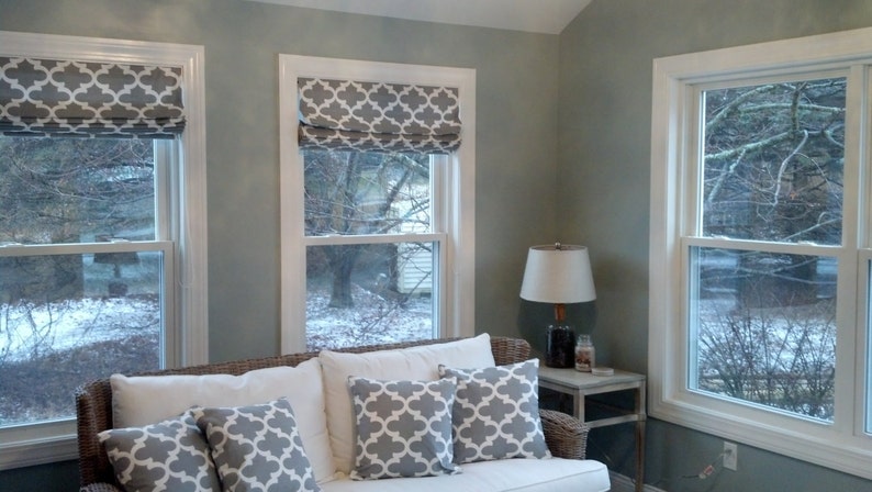 Peut inclure: Un salon avec un canap&eacute; blanc et des coussins &agrave; motifs gris. Il y a trois fen&ecirc;tres avec des encadrements blancs et des stores romains &agrave; motifs gris. Une lampe se trouve sur une table d'appoint &agrave; c&ocirc;t&eacute; du canap&eacute;.