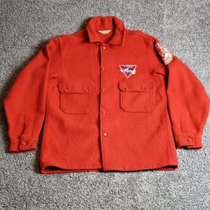 May include: A vintage red wool shirt jacket with a button-down front, two chest pockets, and a collar. The jacket features embroidered patches on the chest and sleeve, with the text "YMCA" visible on the chest patch. The jacket is a classic piece of outerwear.