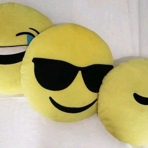 Peut inclure: Trois coussins emoji jaunes. L'un a un visage qui rit avec une larme, un autre a des lunettes de soleil et le troisième a un visage endormi avec des symboles Zzz.