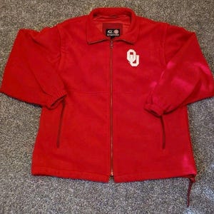 May include: A red fleece jacket with a full zipper closure and a small white logo on the chest. The jacket has a stand-up collar, two front pockets, and elastic cuffs. The jacket is a solid red color.