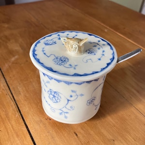 Peut inclure: Un sucrier en céramique blanche avec un motif floral bleu et une petite poignée en forme d'oiseau sur le couvercle. Le bol a un bord bleu assorti et une petite cuillère. Le bol est sur une surface en bois.