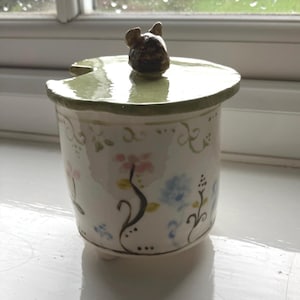 Puede incluir: Jarra de cerámica pintada a mano con tapa. La jarra es blanca con un diseño floral en rosa y azul. La tapa es verde claro con un pequeño asa marrón con forma de animal. La jarra tiene forma cilíndrica.