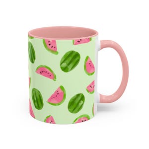Pode incluir: Caneca de cerâmica branca com interior e alça rosa. O exterior apresenta um padrão repetido de fatias e melancias inteiras verdes sobre um fundo verde claro. Um acessório de cozinha divertido e colorido.