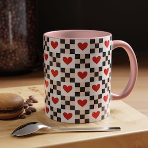 May include: A ceramic coffee mug with a pink interior and handle. The exterior features a pattern of red hearts on a black and white checkered background. The mug sits on a wooden surface with a spoon and a pastry.