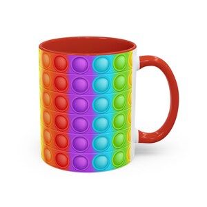 May include: A white ceramic mug with a red interior and handle. The exterior features a colorful, rainbow-colored pattern of circular, raised bubbles, reminiscent of a pop-it toy. The mug is designed for beverages.