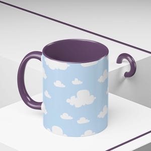 May include: A ceramic mug with a light blue background and white cloud pattern. The interior and handle are a deep purple color. The mug is designed for hot or cold beverages.