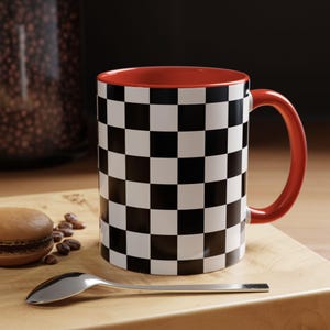 May include: A ceramic coffee mug with a black and white checkered pattern on the exterior. The interior and handle are red. The mug is on a wooden surface with a spoon and a pastry.