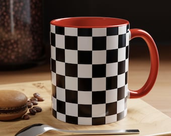 May include: A ceramic coffee mug with a black and white checkered pattern on the exterior. The interior and handle are red. The mug is on a wooden surface with a spoon and a pastry.