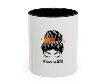 Puede incluir: Taza de cerámica blanca con interior negro. La taza presenta una ilustración negra de la cara de una mujer con los ojos cerrados y un peinado de moño adornado con hojas naranjas. El texto "#momlife" está debajo de la cara.