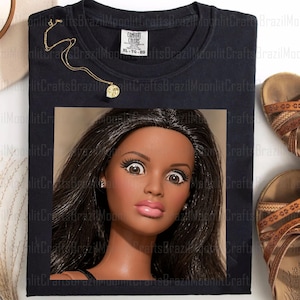 May include: Black t-shirt with a doll face graphic. The shirt has a Comfort Colors tag, size XL-TG-EG. A gold necklace with a pendant is on the shirt. Brown sandals are on the right side of the shirt.