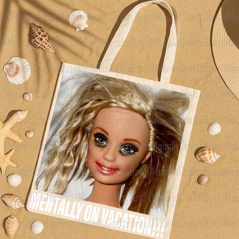 May include: A beige tote bag featuring a doll's face with smudged makeup. The text "MENTALLY ON VACATION!!!" is printed in white. The bag is surrounded by seashells and a straw hat on a sandy surface.