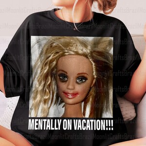 May include: Black t-shirt featuring a close-up of a doll's face with messy blonde hair and dark eye makeup. The text "MENTALLY ON VACATION!!!" is printed in white below the image. The shirt is a casual wear item.