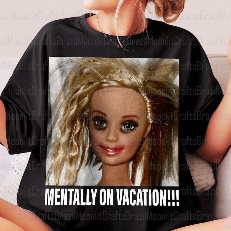 May include: Black t-shirt featuring a close-up of a doll's face with messy blonde hair and dark eye makeup. The text "MENTALLY ON VACATION!!!" is printed in white below the image. The shirt is a casual wear item.