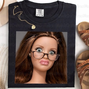 Puede incluir: Camiseta negra con un gráfico de cara de muñeca con gafas y haciendo una mueca divertida. También se ven un collar dorado y sandalias marrones. La camiseta tiene una etiqueta Comfort Colors, talla XL-TG-EG.