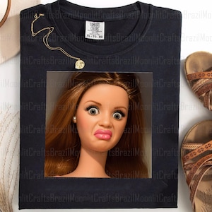 Puede incluir: Camiseta negra XL Comfort Colors con un gráfico de cara de muñeca con expresión de sorpresa. También se ven un collar dorado y sandalias marrones. La camiseta tiene la etiqueta Comfort Colors.