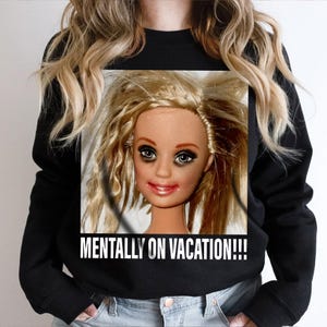 May include: Black sweatshirt featuring a doll face with messy blonde hair, dark eye makeup, and red lipstick. The text "MENTALLY ON VACATION!!!" is printed in white below the doll's face.