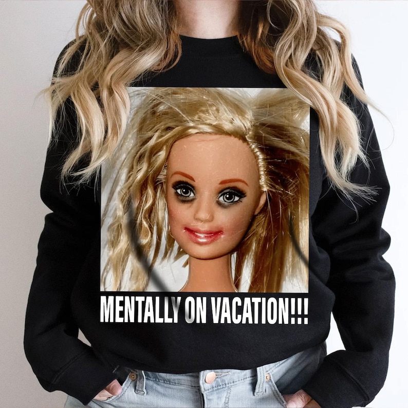 May include: Black sweatshirt featuring a doll face with messy blonde hair, dark eye makeup, and red lipstick. The text "MENTALLY ON VACATION!!!" is printed in white below the doll's face.