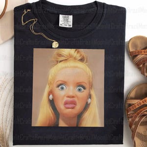 Puede incluir: Camiseta negra con un gráfico cuadrado de una mujer rubia con los ojos muy abiertos y una expresión de sorpresa. La camiseta tiene una etiqueta Comfort Colors. También se ven un collar dorado y sandalias marrones.