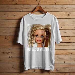 May include: White t-shirt featuring a distressed doll face graphic with the text "MENTALLY ON VACATION!!!" in white. The doll has blonde hair and makeup. The shirt is hanging on a wooden hanger.