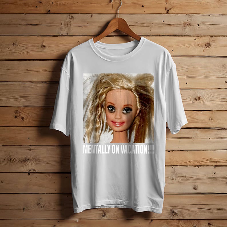 May include: White t-shirt featuring a distressed doll face graphic with the text "MENTALLY ON VACATION!!!" in white. The doll has blonde hair and makeup. The shirt is hanging on a wooden hanger.