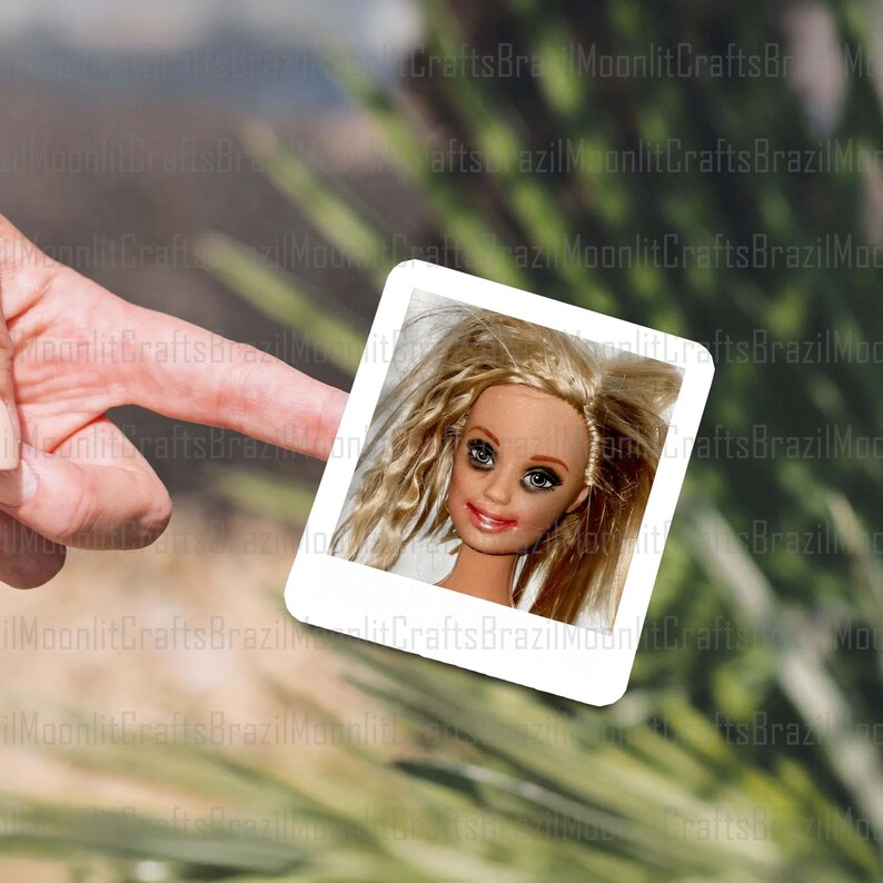 May include: A square photograph featuring a doll's face with blonde hair and blue eyes. The doll has a smiling expression with red lipstick. The photograph is held by a finger against a blurred background of green and brown.