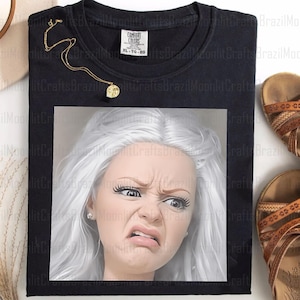 Puede incluir: Camiseta negra Comfort Colors con un gráfico de una mujer con una expresión de disgusto. Un collar dorado con un colgante está sobre la camiseta. Se ven sandalias marrones a la derecha.