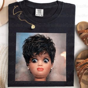 Puede incluir: Camiseta negra con la cara de una muñeca con ojos muy abiertos y expresión de sorpresa. También se ven un collar dorado y sandalias marrones. La camiseta está etiquetada XL-TG-EG.