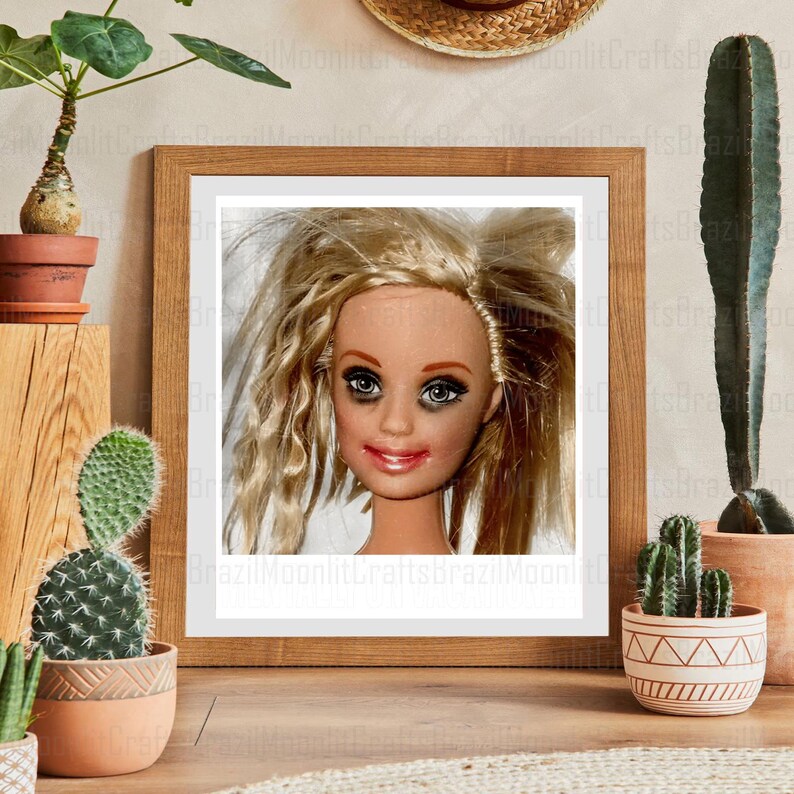 May include: A framed print featuring a doll's head with blonde hair, dark eye makeup, and red lipstick. The doll is set against a white background within a wooden frame. The image is surrounded by potted cacti and a straw hat.