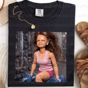 Pode incluir: Camiseta preta com uma imagem emoldurada de uma boneca levantando halteres azuis. A boneca usa um top e shorts rosa, com uma expressão de esforço. Um colar de ouro e sandálias marrons também são visíveis.