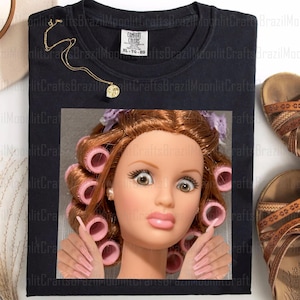 May include: Black XL-TG-EG Comfort Colors t-shirt featuring a doll's face with pink curlers and long pink nails. A gold necklace with a pendant is on the shirt. Brown sandals are on the right.