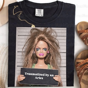 May include: Black t-shirt featuring a doll with smeared makeup holding a sign that reads "Traumatized by an Aries." A gold necklace and brown sandals are also visible. The shirt is labeled XL-TG-EG.