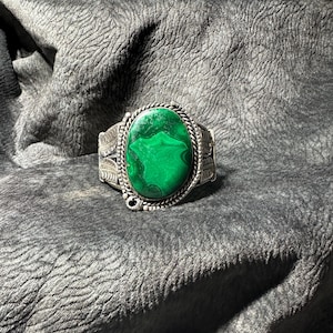 May include: A silver cuff bracelet featuring a large, oval malachite stone in vivid green. The stone is set within an intricate, decorative frame. Other rings and bracelets with different stones are visible, indicating a jewellery collection.