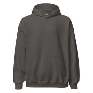 May include: A dark gray hoodie with a front pocket and a hood. The text "RUSTIG AAN" is printed in a lighter gray color on the upper chest area. The hoodie is made of a soft material.