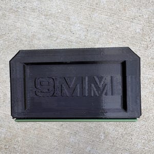 May include: A rectangular black plastic block with the text 9MM embossed in the center. The object features a layered, 3D-printed texture on its surface and a thin, light green horizontal accent line visible along the bottom edge of the item.