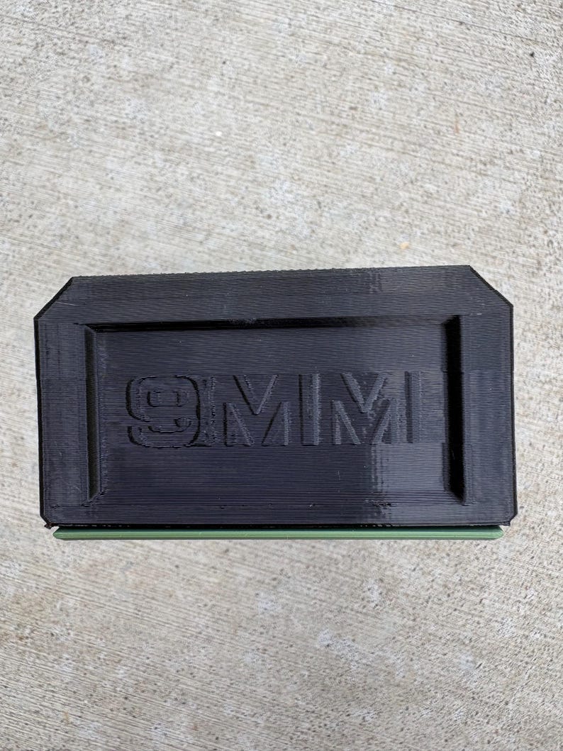 May include: A rectangular black plastic block with the text 9MM embossed in the center. The object features a layered, 3D-printed texture on its surface and a thin, light green horizontal accent line visible along the bottom edge of the item.