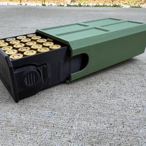 May include: A rectangular storage container with a matte olive green outer sleeve and a black inner drawer. The black drawer is partially extended, revealing a grid of brass-colored ammunition cartridges arranged in rows inside the storage unit.
