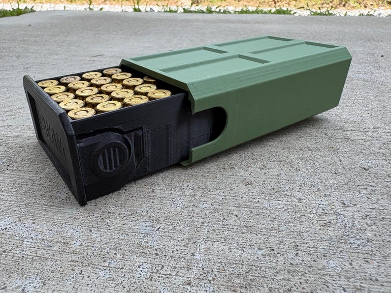 May include: A rectangular storage container with a matte olive green outer sleeve and a black inner drawer. The black drawer is partially extended, revealing a grid of brass-colored ammunition cartridges arranged in rows inside the storage unit.