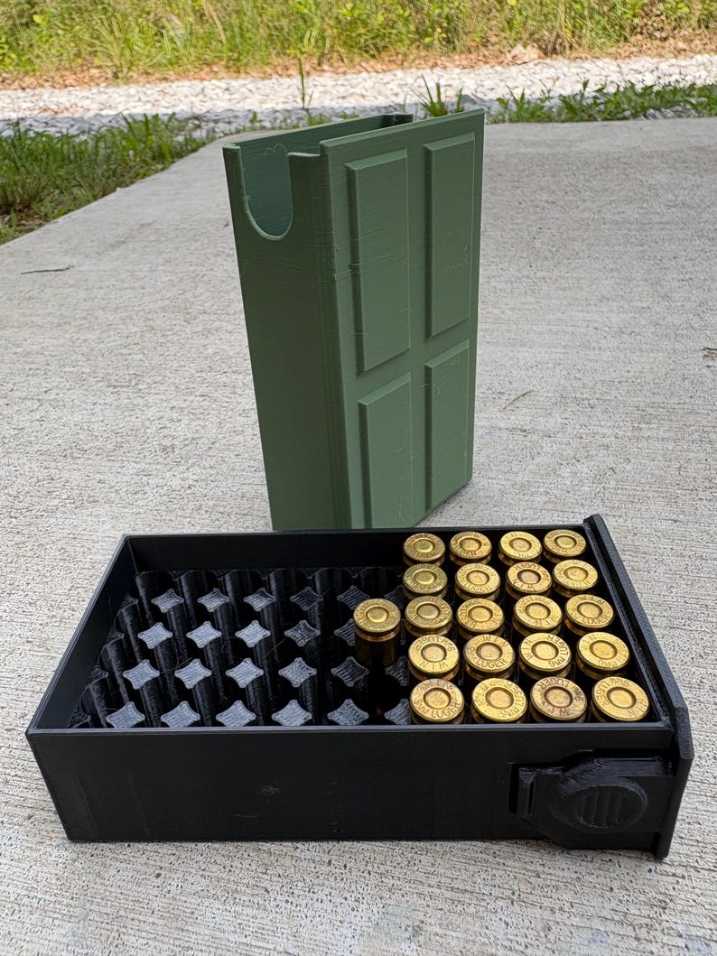 May include: A matte black rectangular ammunition storage tray with a grid of circular slots, partially filled with brass-cased 9mm Luger cartridges. Behind the tray stands an olive green, rectangular, door-shaped storage container.
