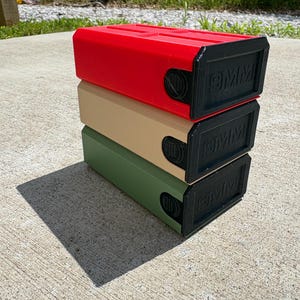 May include: A stack of three rectangular storage containers in red, tan, and olive green. Each container features a black front panel with the embossed text 9MM and a circular side dial mechanism. The units are stacked vertically on a concrete surface.