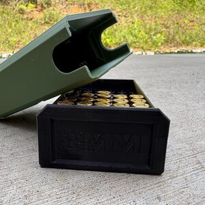 May include: An olive green plastic storage case with a removable lid, containing multiple brass-colored cartridges. The black front panel of the base features the embossed text 9MM, indicating the caliber of the ammunition stored inside the container.