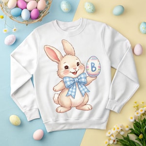 Op de afbeelding: Witte sweatshirt met een cartoon konijn met een blauwe strik, met een versierd paasei met de letter "B". Het ontwerp is gecentreerd op de voorkant. Pasen thema.